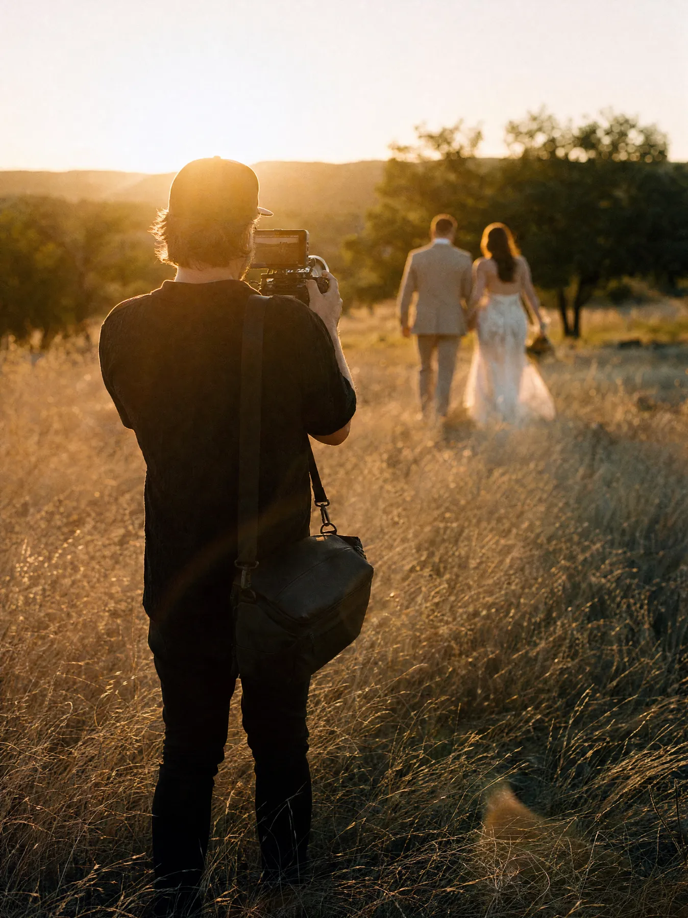 Cinematographer filming a couple walking through tall grass at sunset