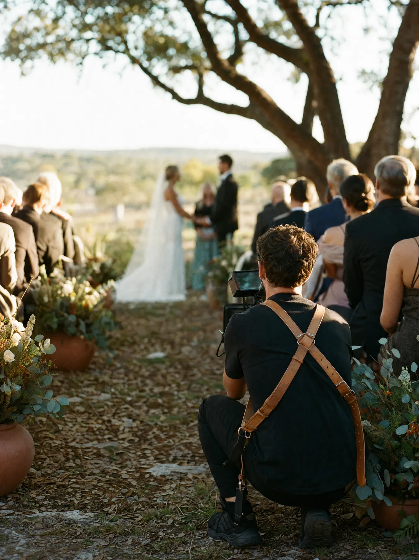 Cinematographer crouched in the aisle filming an outdoor Hill Country ceremony